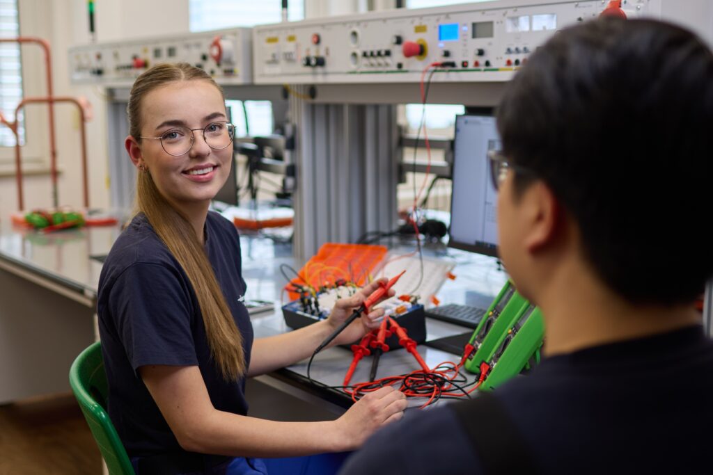 Junge Frau mit Brille lächelt in einem Elektroniklabor, hält Messkabel und arbeitet an Schaltbrett neben Messgeräten.