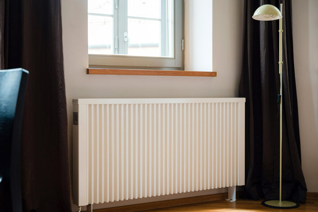 Weißer Wandheizkörper mit vertikalen Rippen unter einem Fenster, rechts schwarzer Vorhang und gelbe Stehlampe.