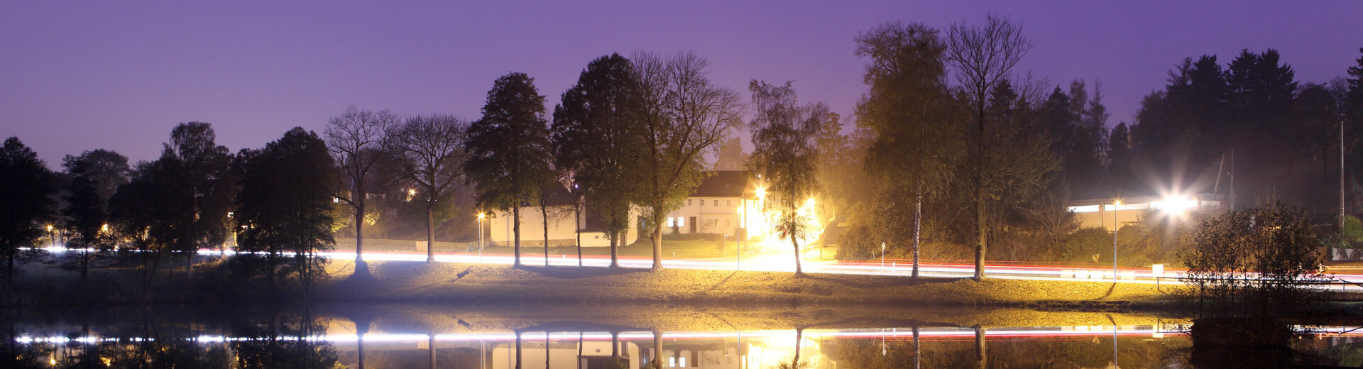 Uferstraße bei Dämmerung mit beleuchteten Bäumen und Häusern, Lichtspuren vorbeifahrender Autos und spiegelnder Seeoberfläche.