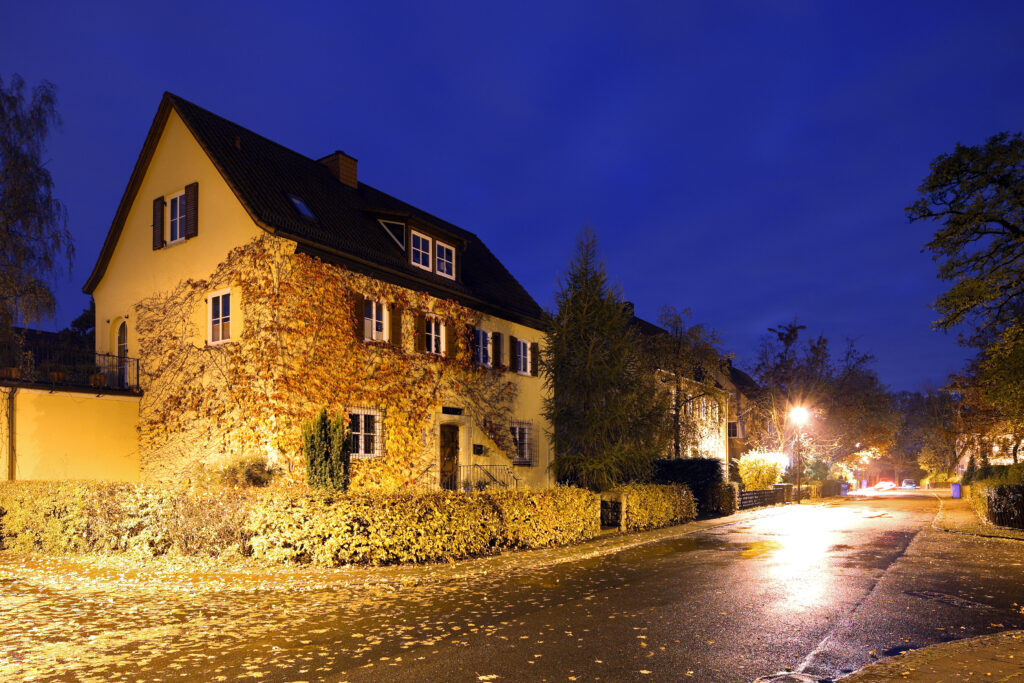 Gelb beleuchtetes Wohnhaus mit Efeu an der Fassade neben einer nassen, herbstlichen Straße bei Nacht